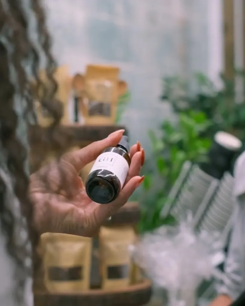 A hand holding a small amber product bottle in front of shelves of kraft paper pouches and plants at a pop-up market